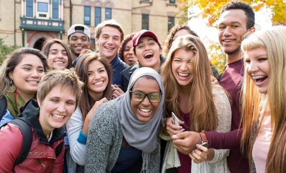 A group of smiling, laughing  students gathered in front of Old Main and leaning together for the picture