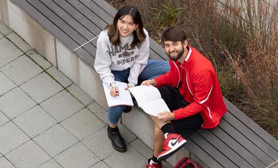 Two  students sitting on a bench outside looking up at the camera smiling