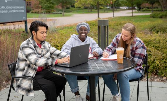Three undergraduate students sitting outside at table on  campus, one with laptop 