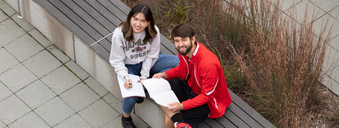Two  students sitting outside on campus with books and smiling up at the camera